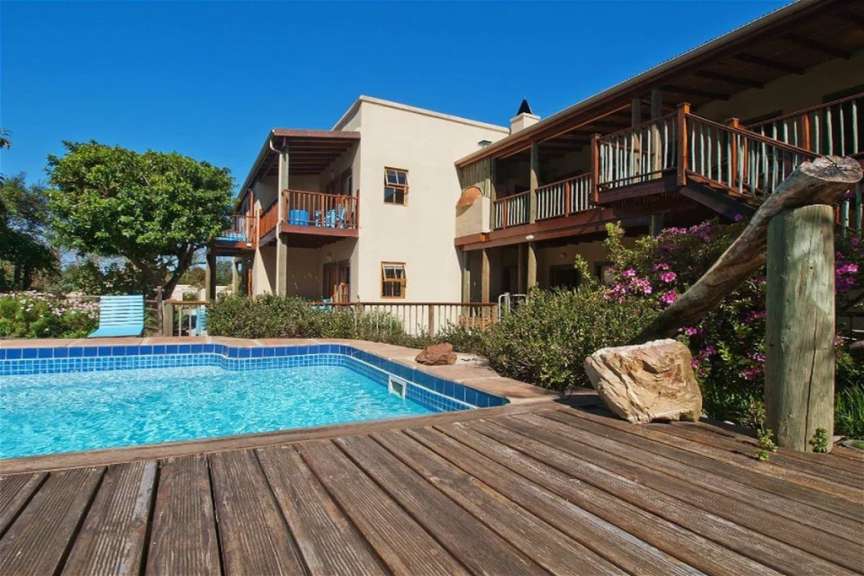 Ein Schwimmbad und eine Holzterrasse grenzen an ein mehrstöckiges Gebäude mit Balkonen, umgeben von üppiger Vegetation und Bäumen unter einem klaren blauen Himmel.