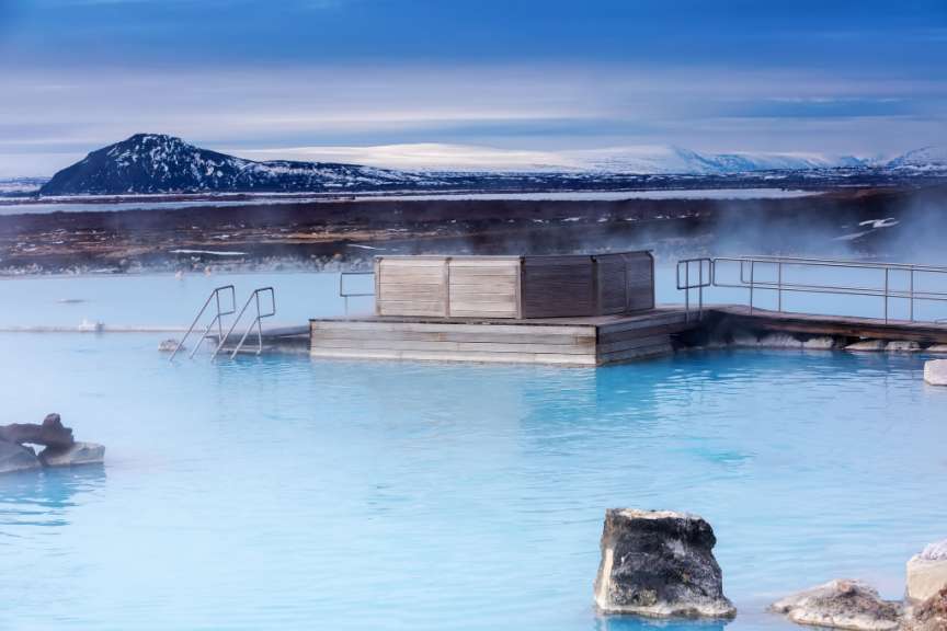 Ein hellblaues Thermalbad mit Dampf und einem hölzernen Steg steht vor einer schneebedeckten Berglandschaft.