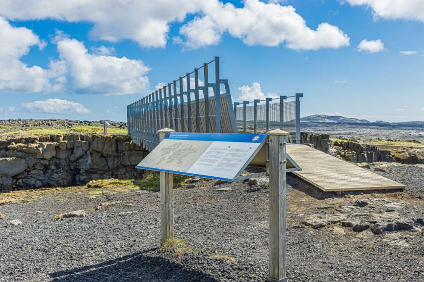 Ein Informationsschild über die tektonischen Platten steht an einem Aussichtspunkt in einer steinigen Landschaft mit einem Holzsteg und einer Mauer aus Felsen.