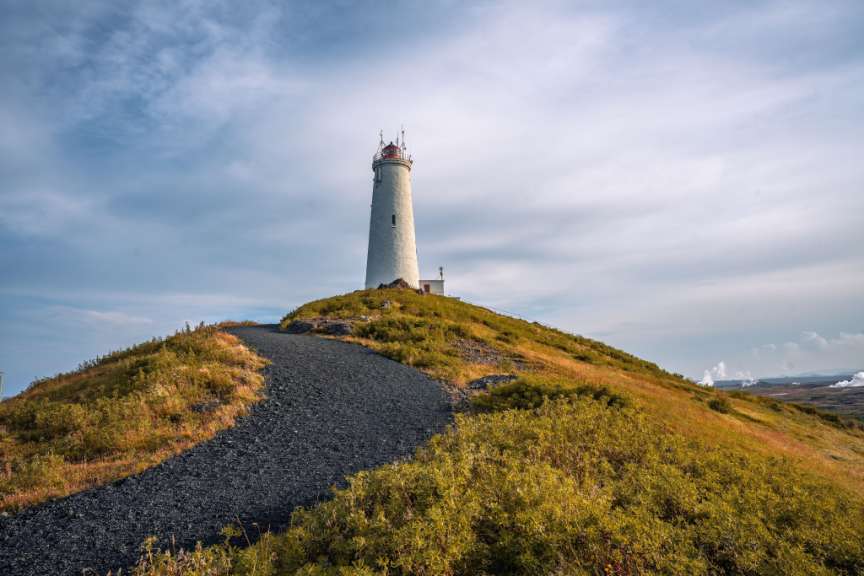 Ein hoher weißer Leuchtturm mit roter Spitze thront auf einem grasbewachsenen Hügel mit einem dunklen Kiesweg, der davor verläuft.