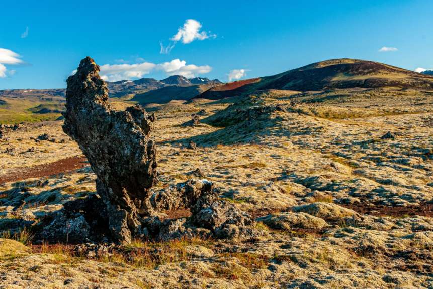 Das Bild zeigt eine raue, moosbedeckte Lavafeldszene mit einem markanten Felsen im Vordergrund, dramatischen Bergen im Hintergrund unter einem strahlend blauen Himmel mit einigen Wolken.