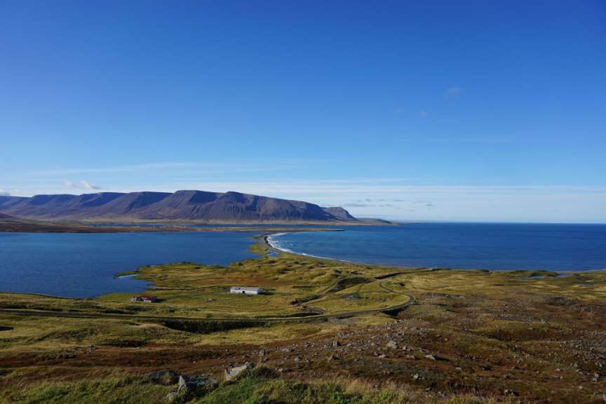 Ein weites Panorama zeigt eine Küstenlandschaft mit blauer See, grünen Hügeln, einer schmalen Landzunge, Bergen im Hintergrund und einem strahlend blauen Himmel.