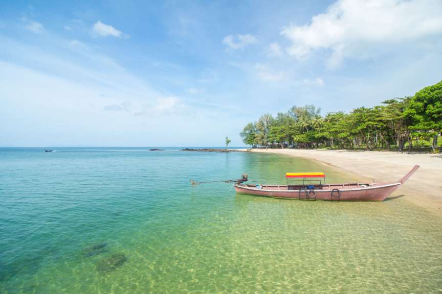 Ein rotes Boot mit gelb-rotem Sonnendach liegt vertäut am Ufer eines tropischen Strandes mit kristallklarem Wasser und üppiger Vegetation im Hintergrund.