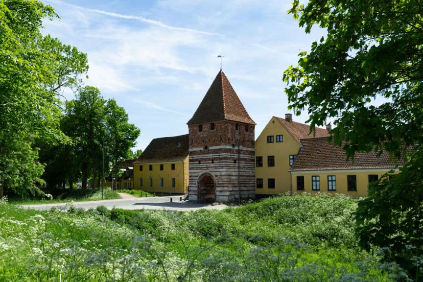 Vor einem blauen Himmel mit weißen Wolken steht eine alte Ziegelsteinturm mit einem roten Ziegeldach, flankiert von gelben Gebäuden mit roten Ziegeldächern, und davor wächst üppiges grünes Gras mit vielen kleinen weißen Blumen.