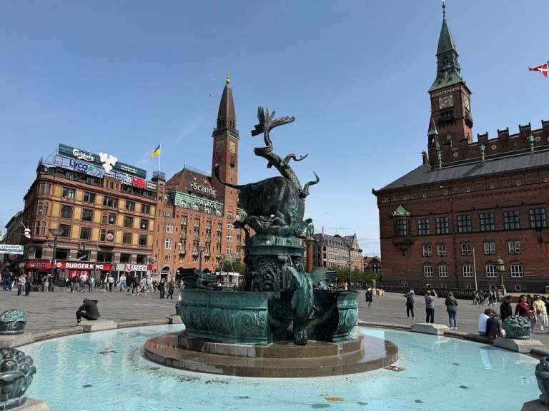 Ein Brunnen auf dem Rathausplatz in Kopenhagen steht vor dem Rathaus und weiteren Gebäuden, während Menschen den Platz beleben.