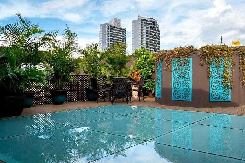 Ein moderner Dachgarten mit Pflanzen, Sitzgelegenheiten und einem Glasboden, der einen Pool darunter zeigt, mit Hochhäusern im Hintergrund unter einem bewölkten Himmel.