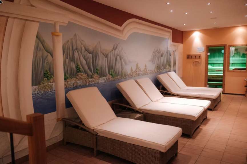 Wellnessbereich mit hellen Liegen und einem großen Bergpanorama-Wandgemälde vor einer Sauna.