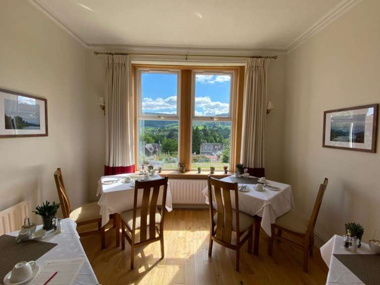 Zwei mit weißen Tischdecken gedeckte Tische mit Gedeck stehen vor einem Fenster mit Blick auf eine grüne Landschaft, im Vordergrund ist ein Tisch mit einem aufgeschlagenen Menü zu sehen.