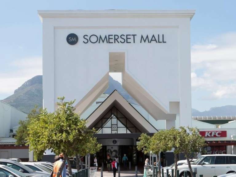 Die weiße Fassade der Somerset Mall in Südafrika zeigt den Haupteingang mit Passanten unter wolkenlosem Himmel.