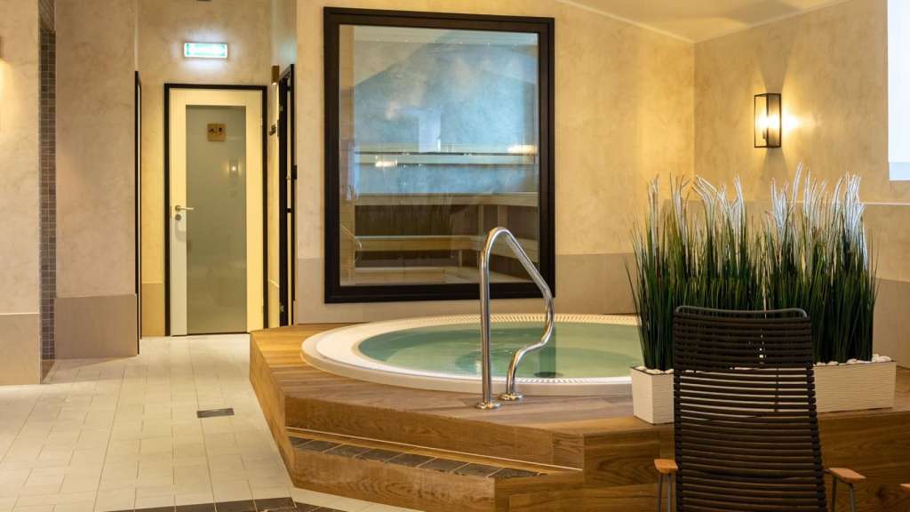 Moderner Wellnessbereich mit einem runden Whirlpool aus Holz auf Fliesenboden und einer Saunakabine im Hintergrund.