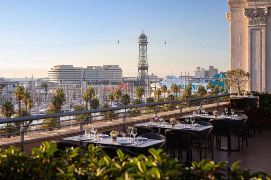 Blick von einer Hotelterrasse am Nachmittag auf den Hafen von Barcelona mit Yachten, Seilbahnturm und Schiffen.