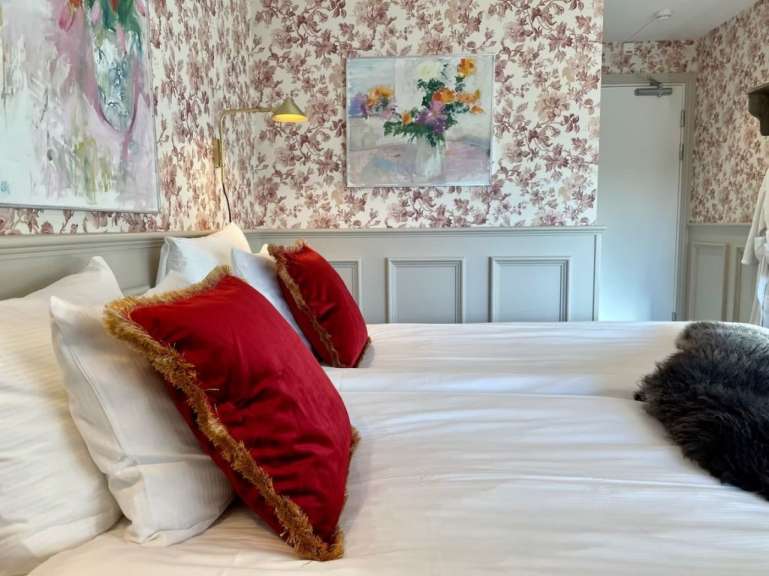 Hotelzimmer in Schweden mit Doppelbett, roten Zierkissen, Gemälden an der Mustertapete und grauem Paneel.