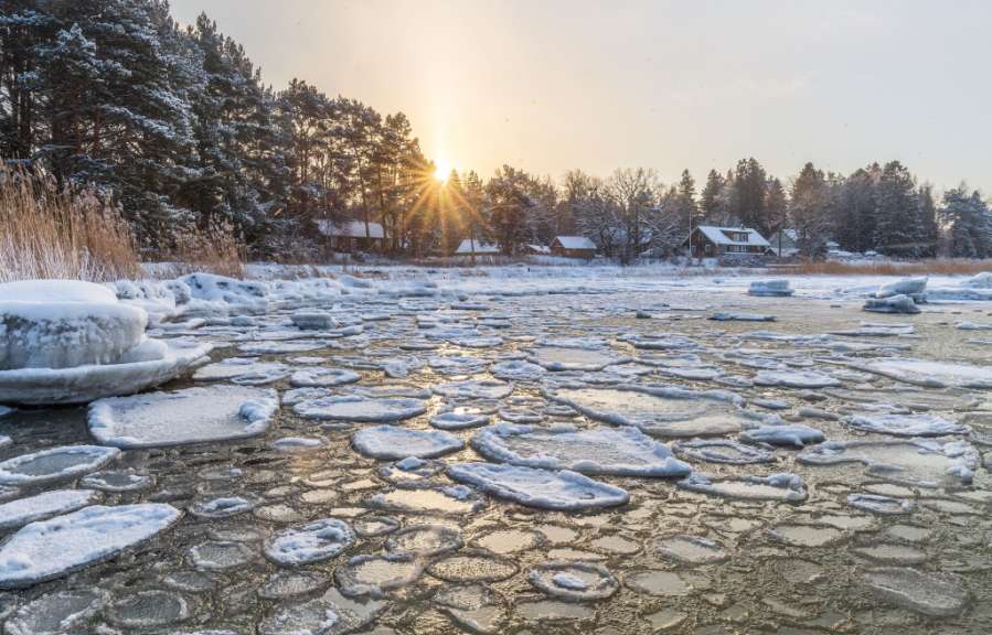 Wintersonne über dem eisbedeckten Meer vor dem estnischen Fischerdorf Altja mit verschneiten Holzhäusern und Bäumen.