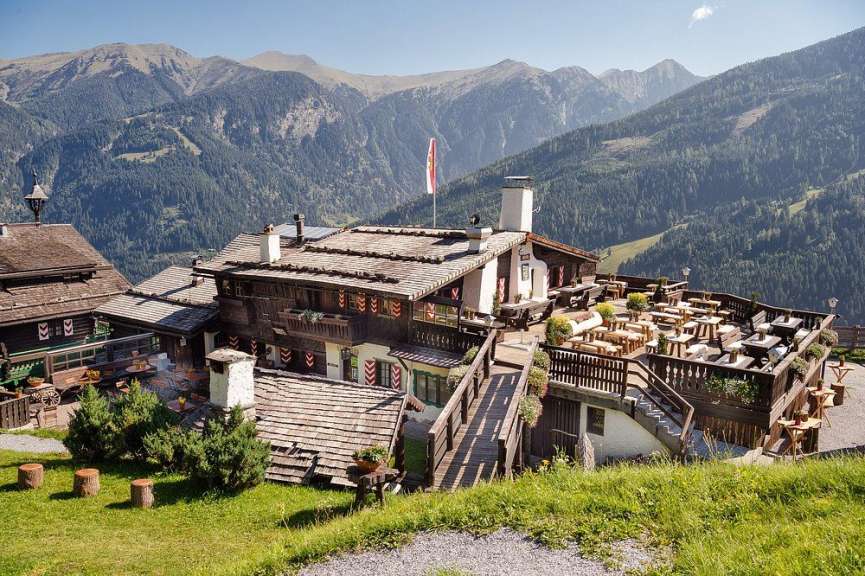 Ein traditionelles Holz-Berghotel in den österreichischen Alpen mit großer Terrasse und Blick auf ein bewaldetes Bergmassiv.