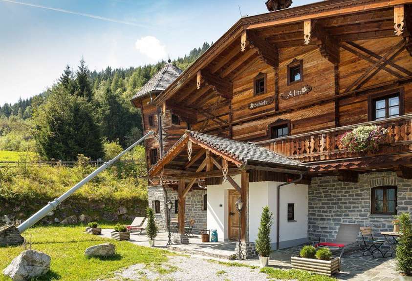 Ein traditionelles Holzhaus mit Steinuntergeschoss und Vordach steht an einem Hang mit Wald in Österreich.