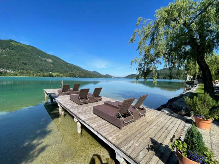 Mehrere Liegen stehen an einem sonnigen Tag auf einem hölzernen Steg am türkisblauen See in Österreich.