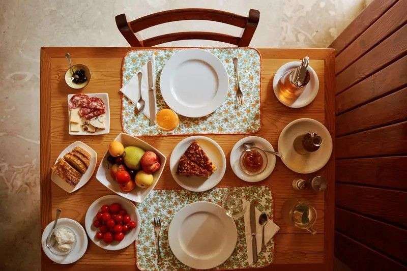 Ein italienischer Frühstückstisch ist mit Tellern, Brot, Aufschnitt, Früchten, Kuchen und Getränken gedeckt.
