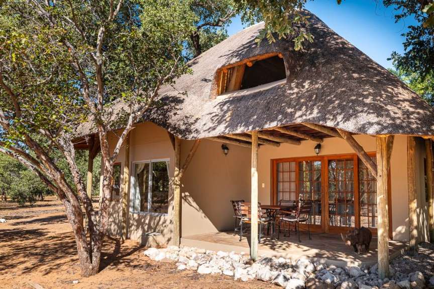 Ein Bungalow mit Reetdach und Terrasse steht am Tag in einer buschreichen Landschaft in Namibia.