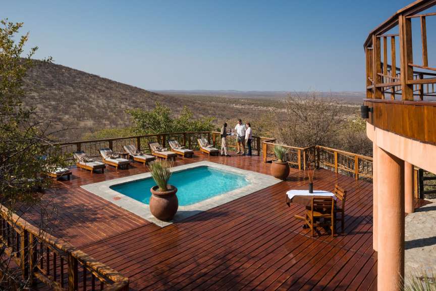 Eine hölzerne Terrasse mit einem Pool und Liegestühlen überblickt eine trockene, hügelige Landschaft unter einem klaren blauen Himmel.