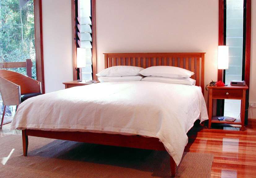 Ein schlicht eingerichtetes Schlafzimmer mit einem weiß bezogenen Bett, Holzboden und einem Fenster mit Jalousien.