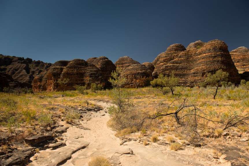 Ein sandiger Weg führt durch eine Wüste mit Felsen im Hintergrund