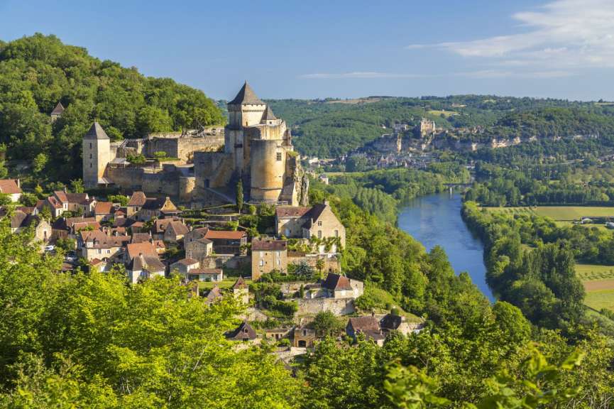 Eine malerische Flusslandschaft mit einer mittelalterlichen Burg und einem Dorf, eingebettet in grüne Hügel unter blauem Himmel.