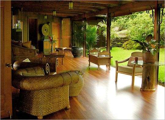 Ein überdachter Sitzbereich mit Holzboden und Möbeln aus Rattan und Holz grenzt an einen grünen Garten mit Steintreppen.