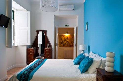 In einem hellen Schlafzimmer steht ein breites Bett vor einer blauen Wand mit einem Nachttisch, auf dem eine weiße Lampe und eine weiße Vase stehen, und einem Spiegel mit einer schmalen Kommode.