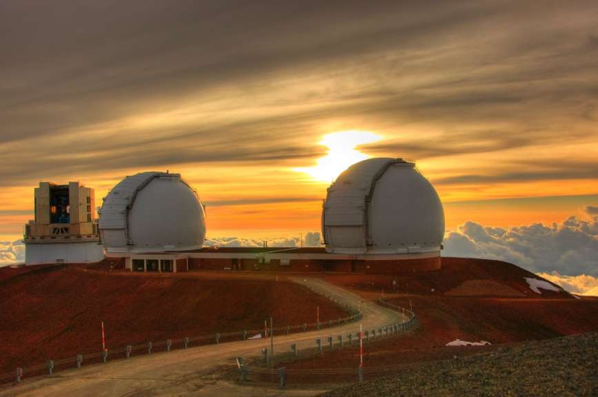 Zwei astronomische Observatorien mit kuppelförmigen Dächern stehen auf einem roten Hügel unter einem dramatischen Sonnenuntergangshimmel.