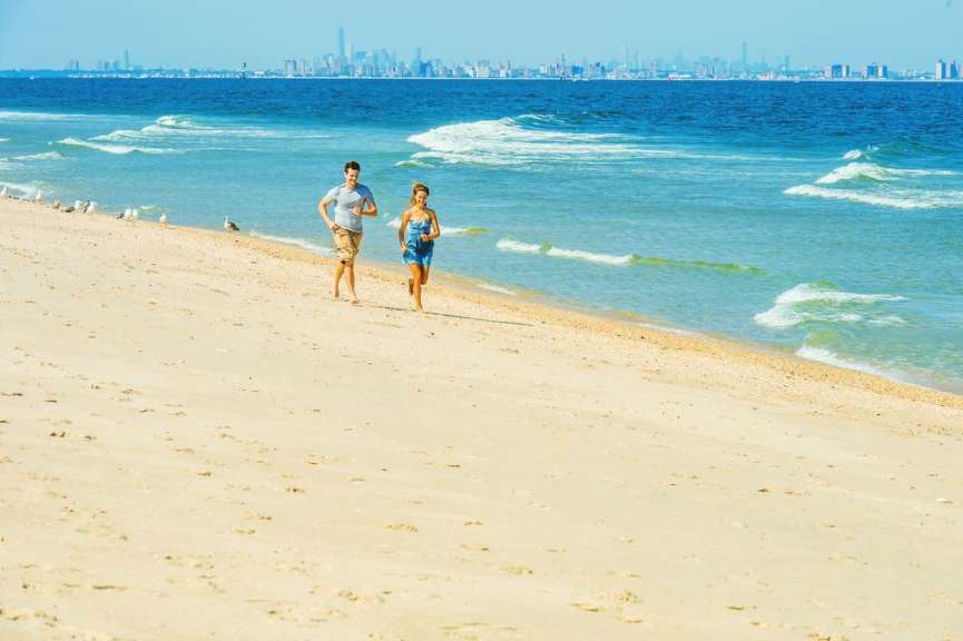 Zwei Menschen joggen am Strand entlang, während im Hintergrund eine Stadt am Horizont zu sehen ist.