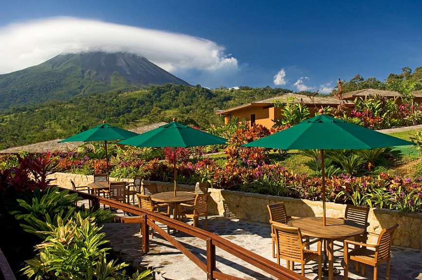 Eine Terrasse mit Tisch und Stühlen und grünen Regenschirmen mit einem Vulkan im Hintergrund