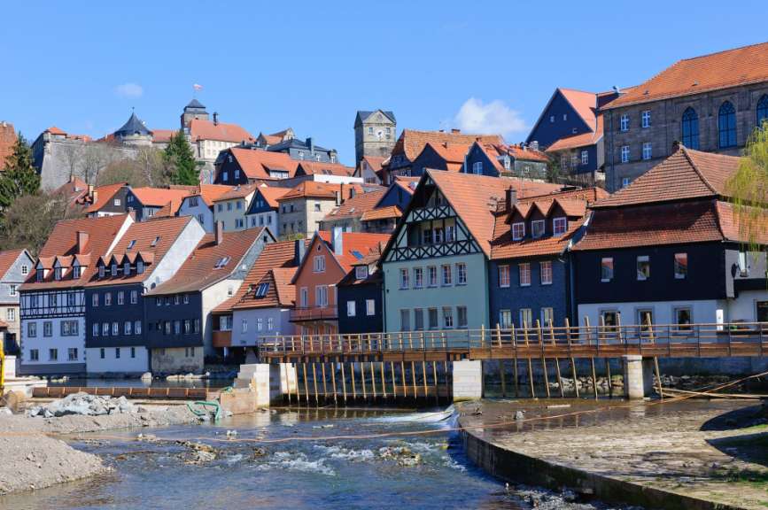 Der Blick über eine Stadt mit vielen Häusern und einem Fluss im Vordergrund zeigt eine Burg und einen Turm im Hintergrund.
