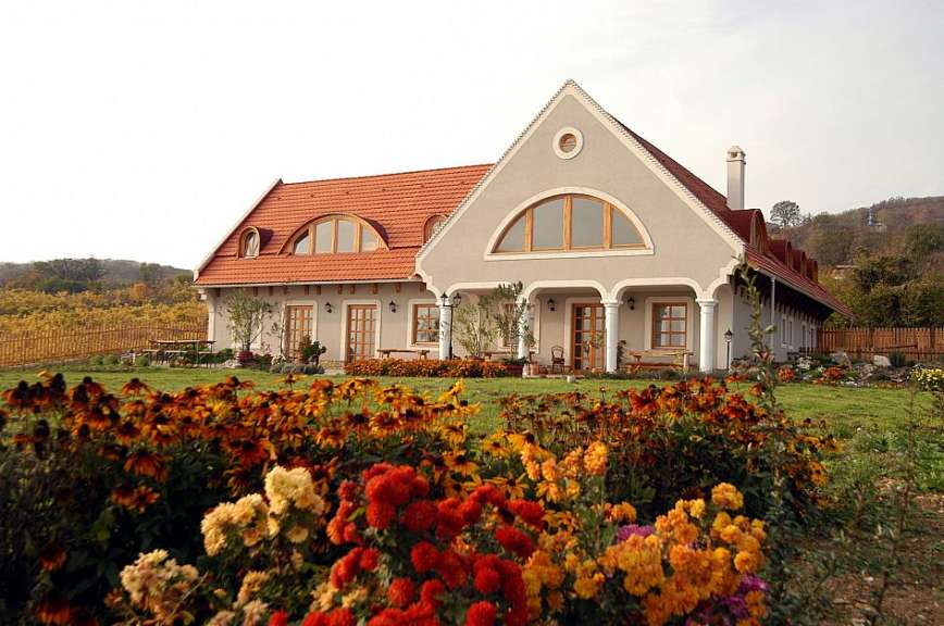 Ein landwirtschaftliches Gebäude mit rotem Ziegeldach und großen Bögenfenstern steht inmitten einer bunten Blumenwiese und eines Gartens mit Holzzaun.