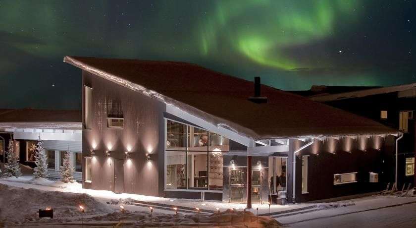 Eine nordische Unterkunft ist im Abendlicht bei einem leuchtenden Nordlicht zu sehen.