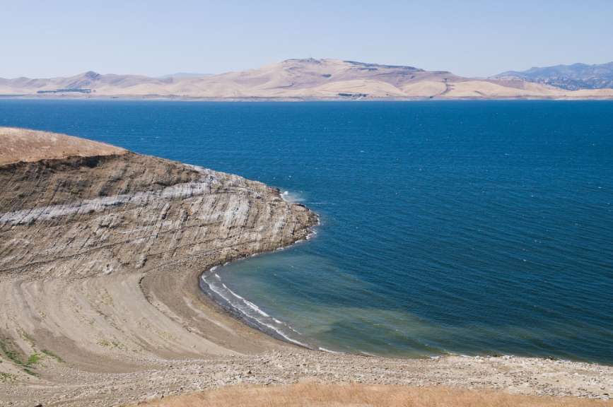 Eine felsige Küste mit geschichteten Gesteinsschichten fällt in ein tiefblaues Wasser, vor dem sich eine hügelige, trockene Landschaft abzeichnet.