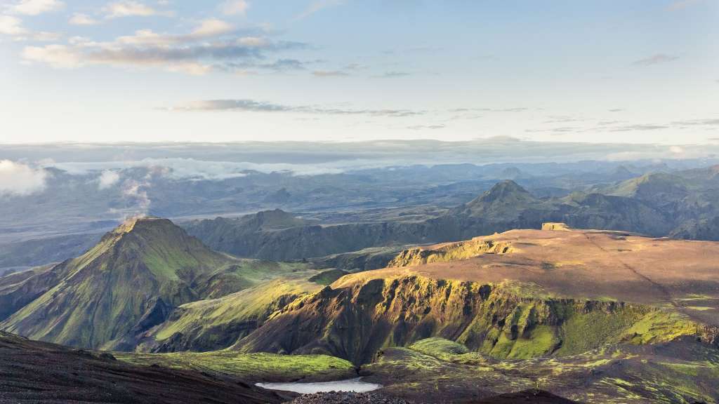 Diese dramatische Landschaftsaufnahme zeigt ein zerklüftetes Gelände mit grünen Hügeln und schluchtenartigen Tälern, die von der Morgensonne beleuchtet werden.