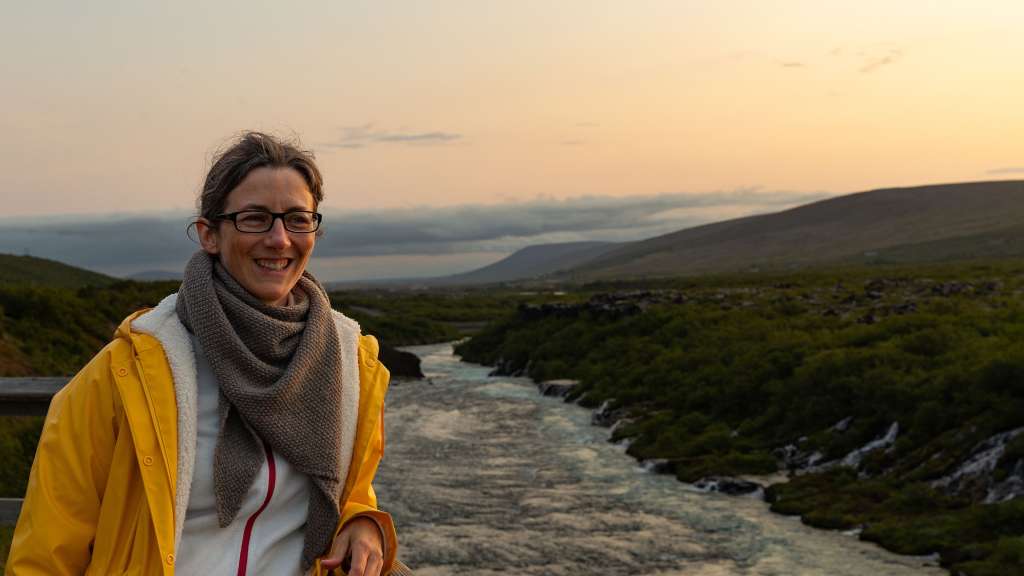 Eine Frau in einem gelben Regenmantel und einem Schal steht an einem Fluss in einer bergigen Landschaft bei Sonnenuntergang.