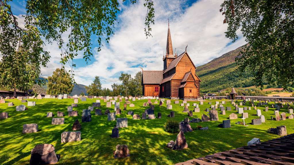 Ein hölzerner Stabkirchenfriedhof mit vielen Grabsteinen, der sich auf einem Hügel hinter einer Mauer befindet, mit einer bewaldeten Bergkulisse im Hintergrund.