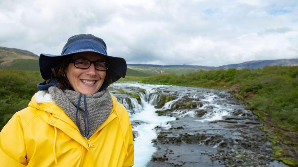 Eine Frau in einem gelben Regenmantel und einem Hut lächelt in die Kamera vor einem breiten, flachen Wasserfall im Freien.