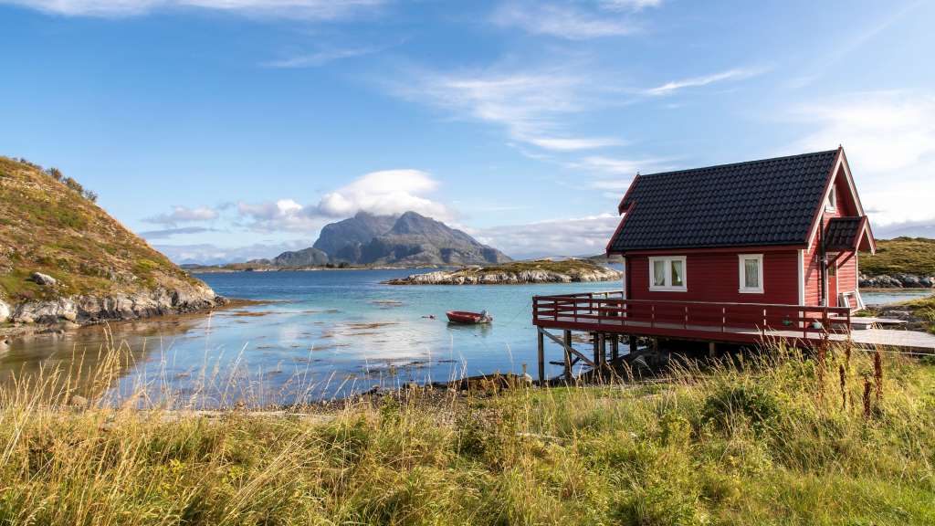 Ein rotes Haus auf Stelzen steht am Ufer eines ruhigen Gewässers mit einem Berg im Hintergrund.