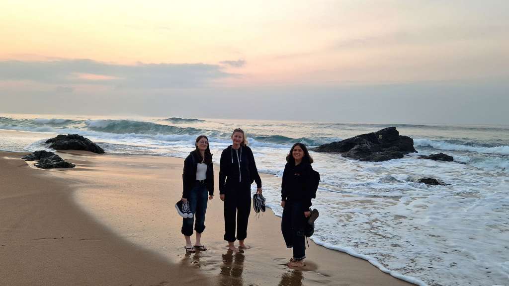 Drei junge Frauen stehen an einem Strand, während die Wellen auf den Sand treffen und die Sonne den Himmel in warmen Tönen färbt.