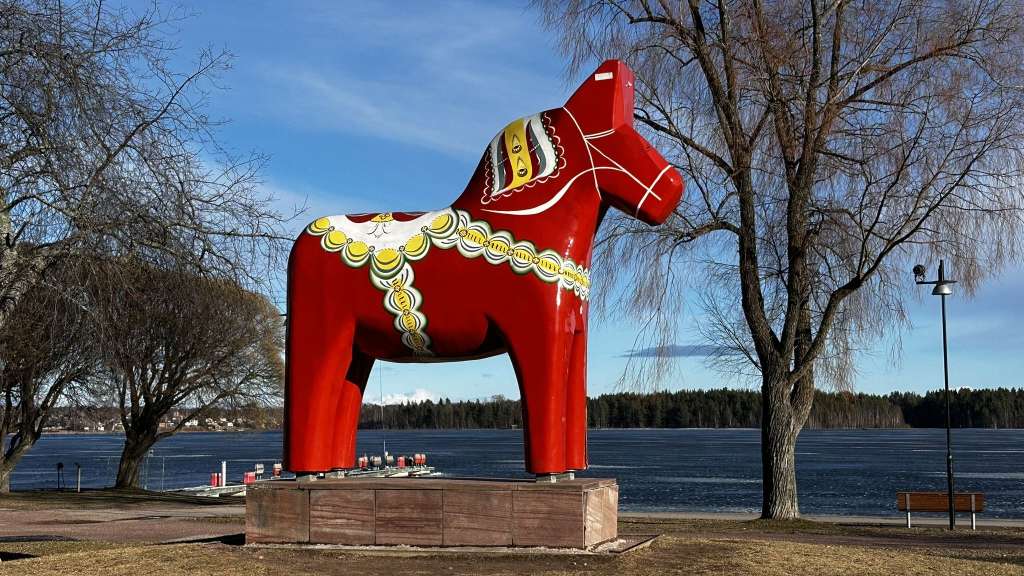 Ein großes, rotes, verziertes Dalapferd steht auf einem Sockel vor einem gefrorenen See mit Bäumen und blauem Himmel.