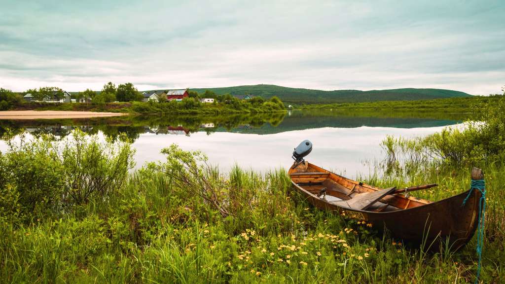 Ein Holzboot mit Außenbordmotor liegt am Ufer eines ruhigen Sees mit einer Ortschaft im Hintergrund unter einem bewölkten Himmel.