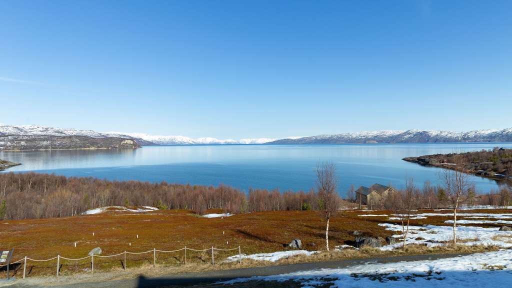 Eine klare, blaue Himmelslandschaft erstreckt sich über ein ruhiges Gewässer, das von schneebedeckten Bergen und einer felsigen Küste gesäumt ist, während dichte, kahle Bäume einen braunen, von Schnee durchsetzten Hügel säumen.