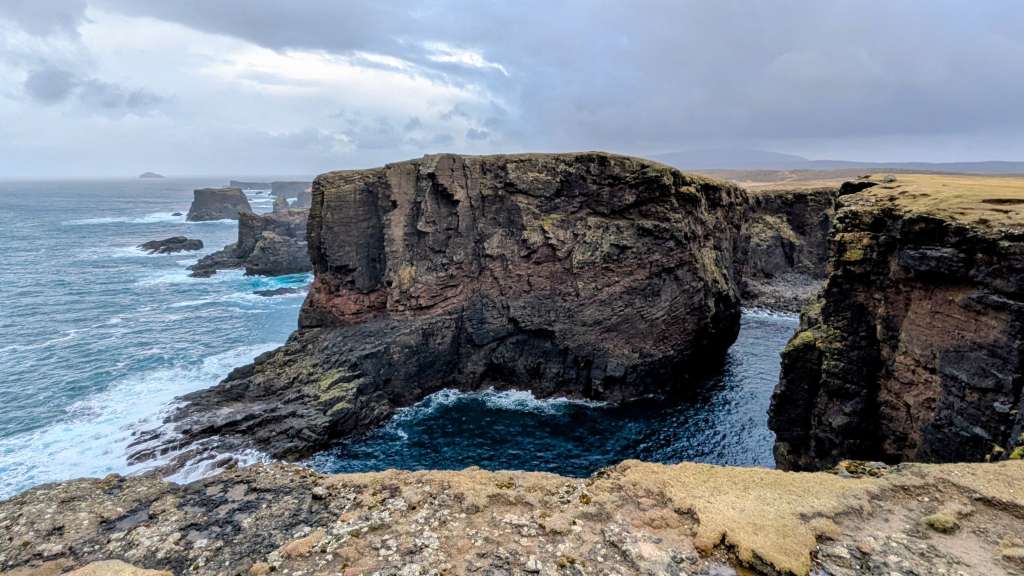 Dieses Bild zeigt die dramatische Küstenlandschaft mit steilen Klippen, felsigen Inseln und aufgewühltem Meer unter einem bewölkten Himmel.