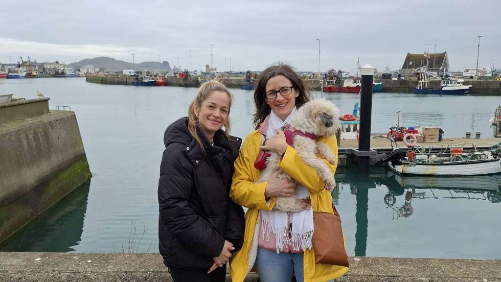 Zwei Frauen stehen mit einem Hund vor einer Hafenkulisse.