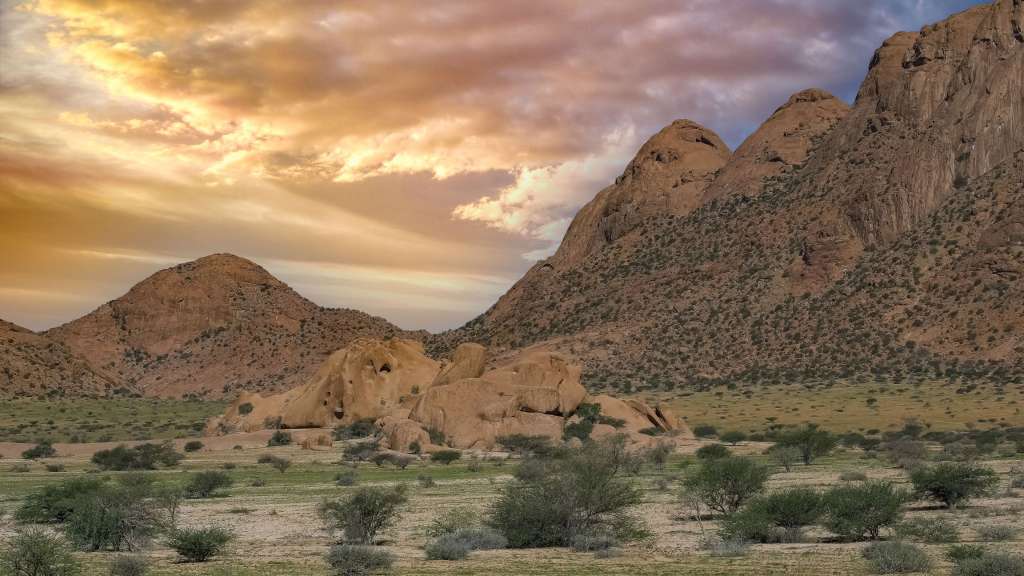 Ein weites, trockenes Tal mit vereinzelten Bäumen und Sträuchern wird von hohen Felsformationen und einem dramatischen Wolkenhimmel dominiert.