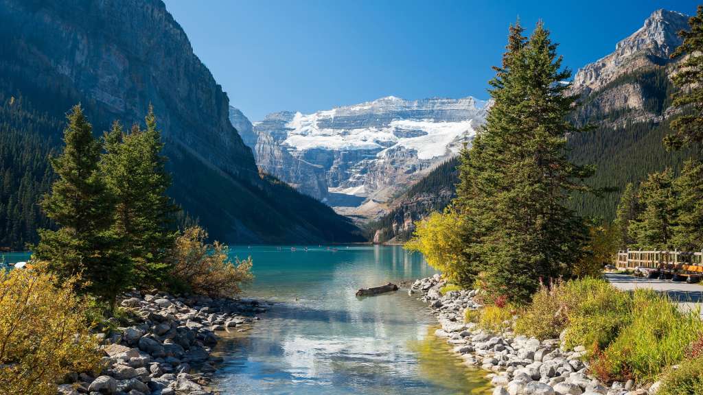 Ein malerischer Blick auf einen türkisfarbenen Bergsee mit schneebedeckten Gipfeln im Hintergrund und felsigen Ufern mit Bäumen und Herbstlaub.