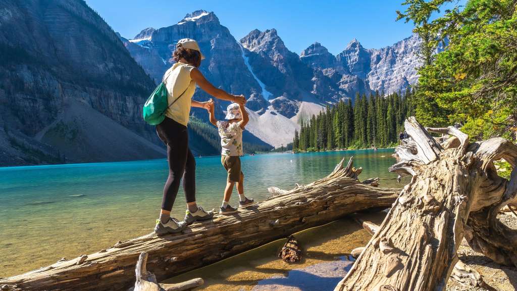Eine Frau und ein Kind balancieren auf einem Baumstamm im klaren Wasser eines Bergsees vor einer imposanten Bergkulisse.