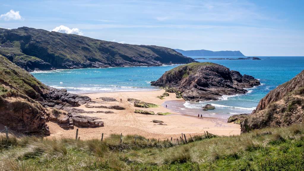 Eine weite Aufnahme einer felsigen Küste mit einem sandigen Strand und dem türkisblauen Meer, über dem ein klarer Himmel mit wenigen Wolken hängt.